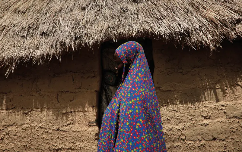 A woman in Cameroun dressed with flower patterned clothes standing in front of a building with straw roof.