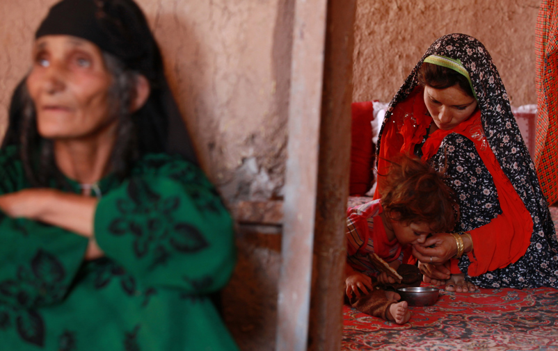 Afghan woman feeds child sitting on the floor.