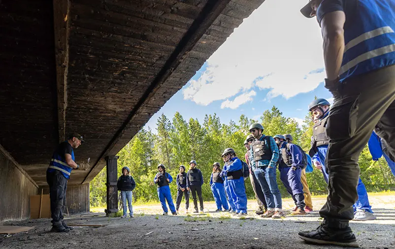 En grupp människor i blå overaller står framför en kursinstruktör i blå väst som står i skuggan under ett skärmtak.