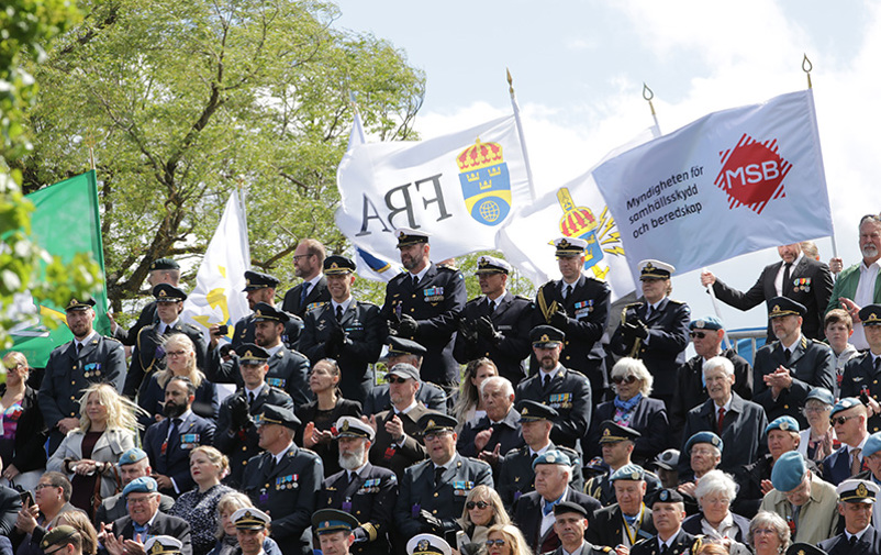 Bild på militära veteraner i uniform som står på en läktare under ett veterandagsfirande. FBA:s logotyp syns på en flagga i bakgrunden.